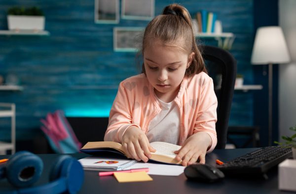 Schoolchild sitting at desk in living room holding school book